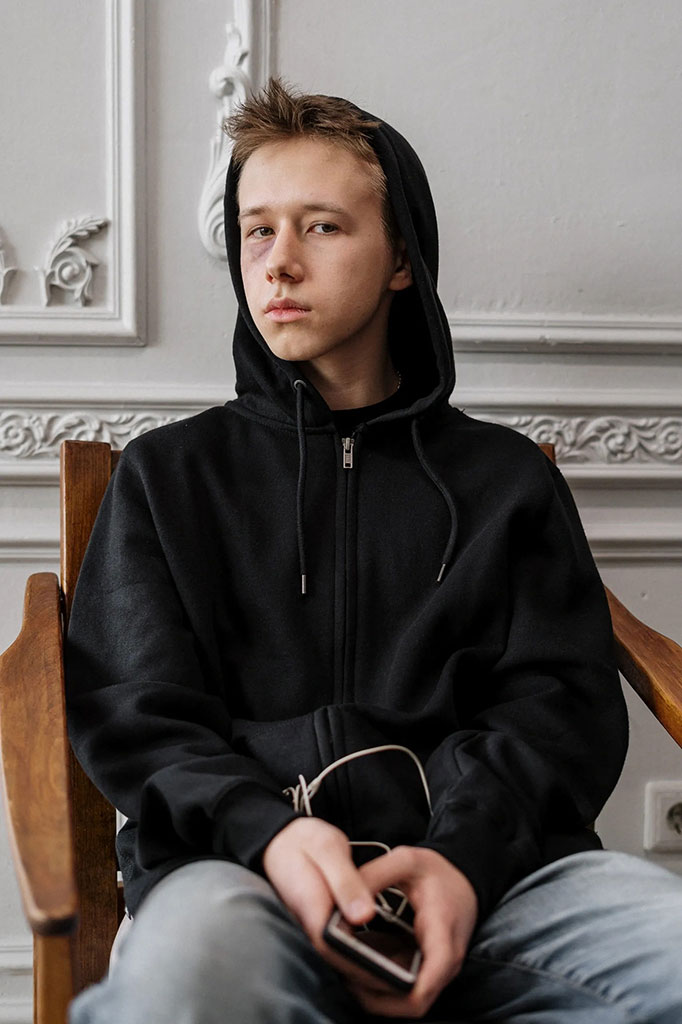 Person in a hoodie sitting with a distant expression, holding a phone, representing struggle with addiction